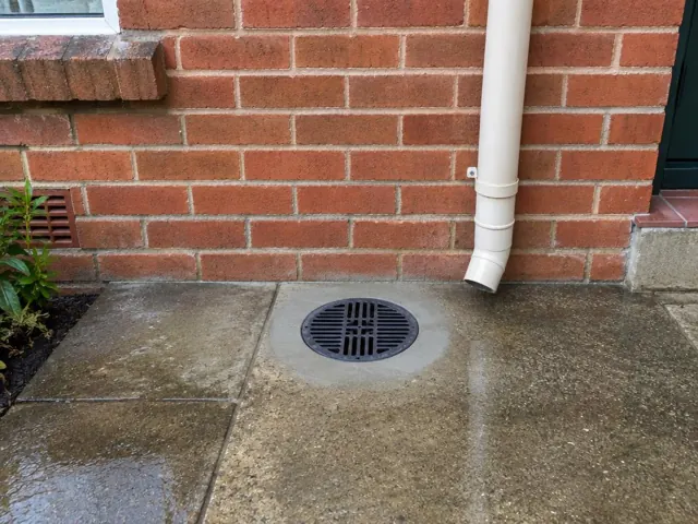Clean outdoor grated drain in a concrete path beside a red brick wall in Kearsley